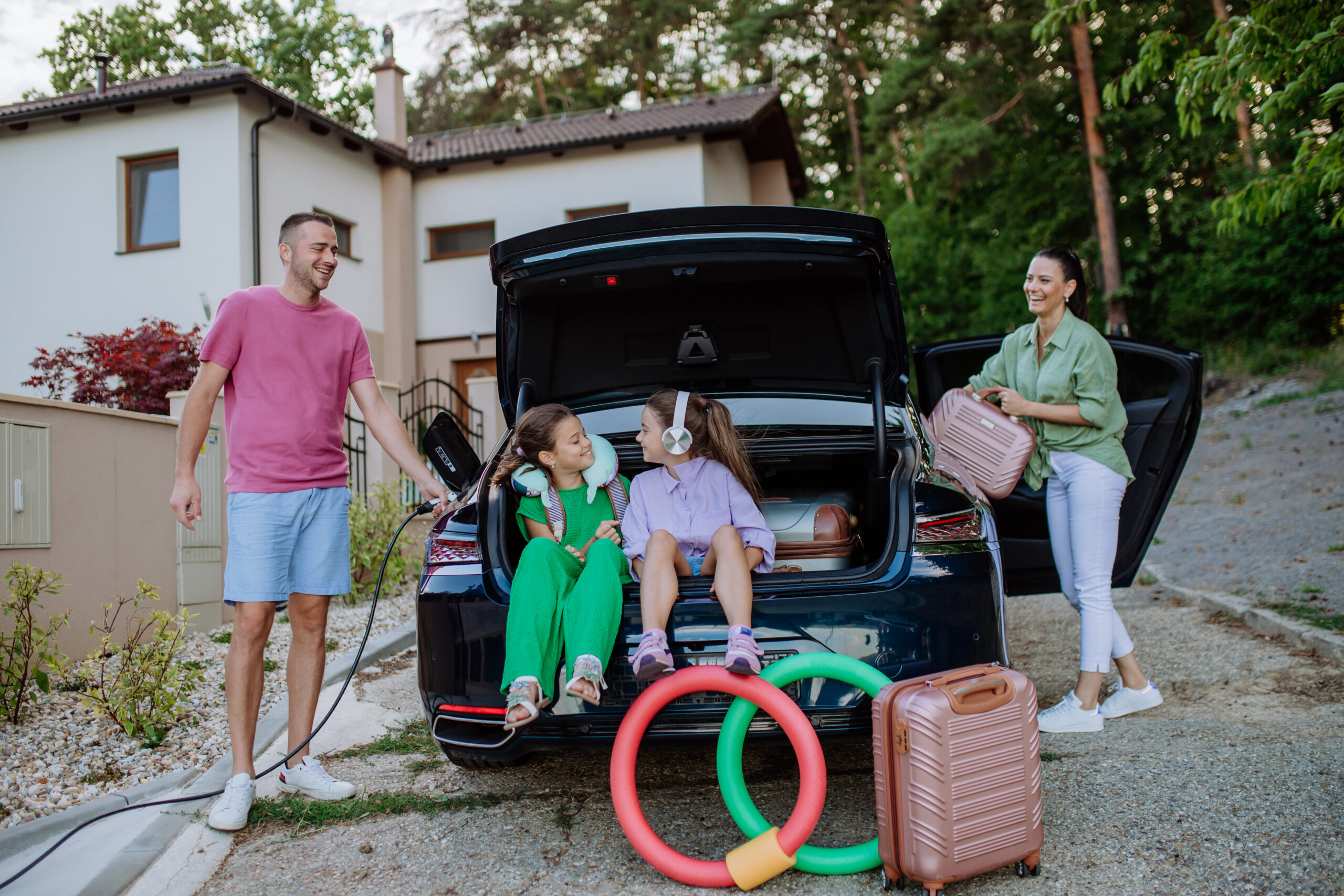 Happy father holding power supply cable and charging their electric car, rest of family putting suitcases in a car trunk, preparating for holidays.